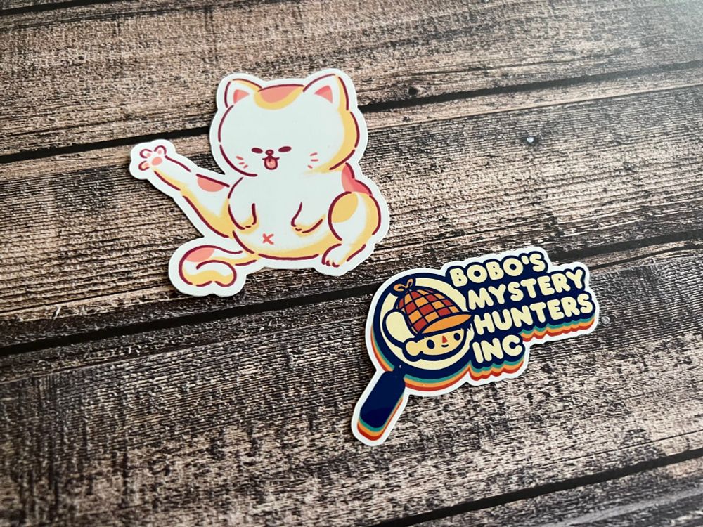 Two die cut stickers. One of a cat with its “chicken leg” out, and one that says “Bobo’s Mystery Hunters Inc” from the video game Mineko’s Night Market. 