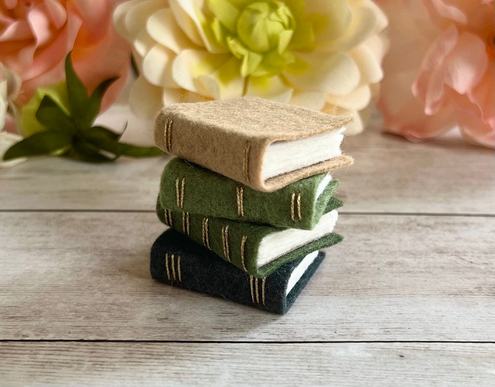 A stack of four mini books made from felt with gold threaded spines in light brown, green, and grey. 