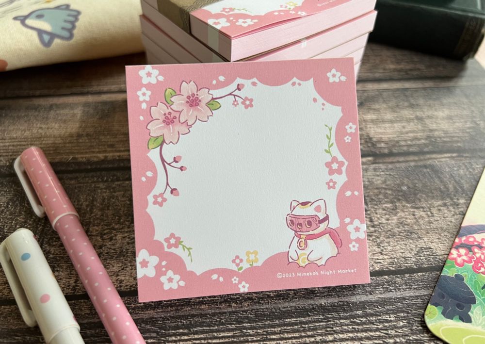 A small, square pink-bordered notepad featuring Nikko from the video game Mineko’s Night Market. 