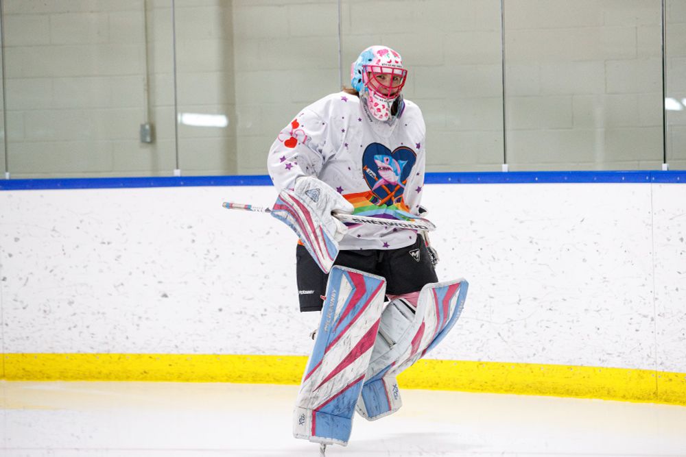 A goalie in white, pink, and blue, skates near the boards, pointing at the camera with the toe of her stick. She's laughing about something unfunny.