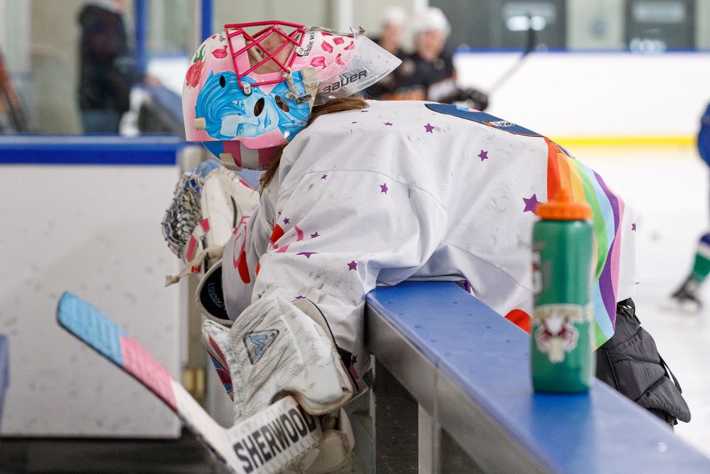 A goalie in white, pink, and blue, stretches her back by folding herself in half over the boards, while looking towards the ceiling. The photographer says that it hurts to look at.