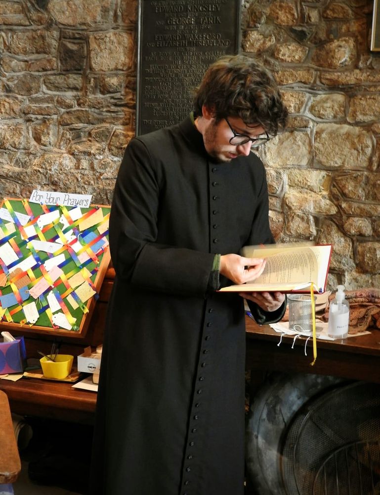 Jay is standing in a black cassock flicking through a prayer book