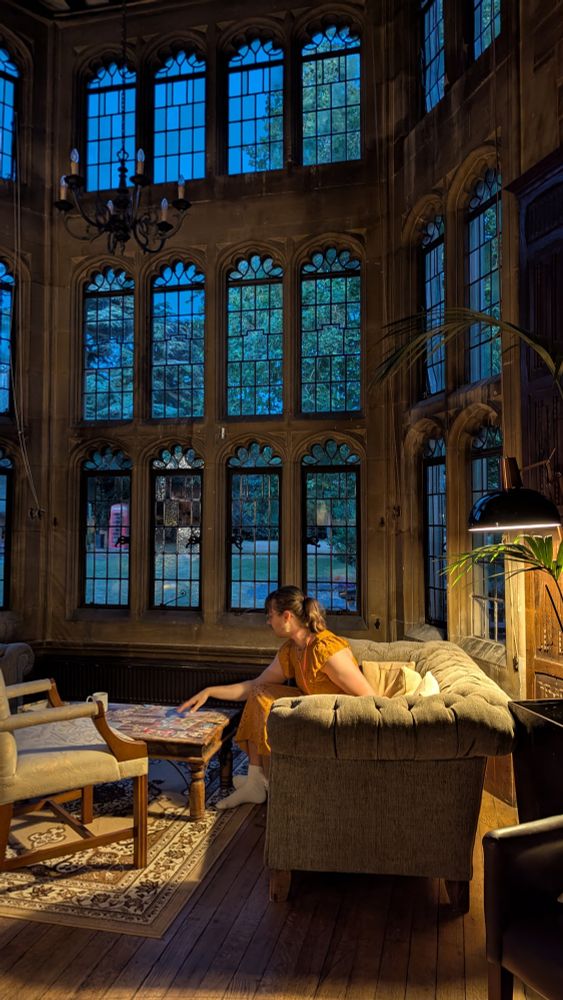 Huw sits on a sofa in a large 17th century window, leaning forward to play a board game on the table before them.