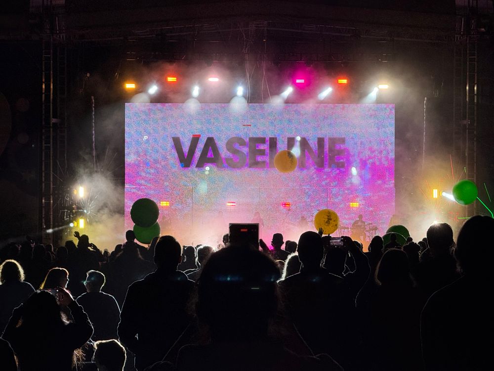 A brightly lit stage with multicolored lights and the word “Vaseline” on a large screen. In the foreground is the silhouettes of concert-goers and beach balls in the air.