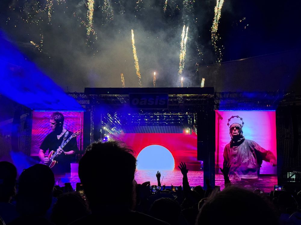 Closing of the Oasis show in Chicago. Noel playing guitar on the left screen, Liam with a tambourine on his head on the right screen, and a sunset on the middle screen. Fireworks are going off over the stage.