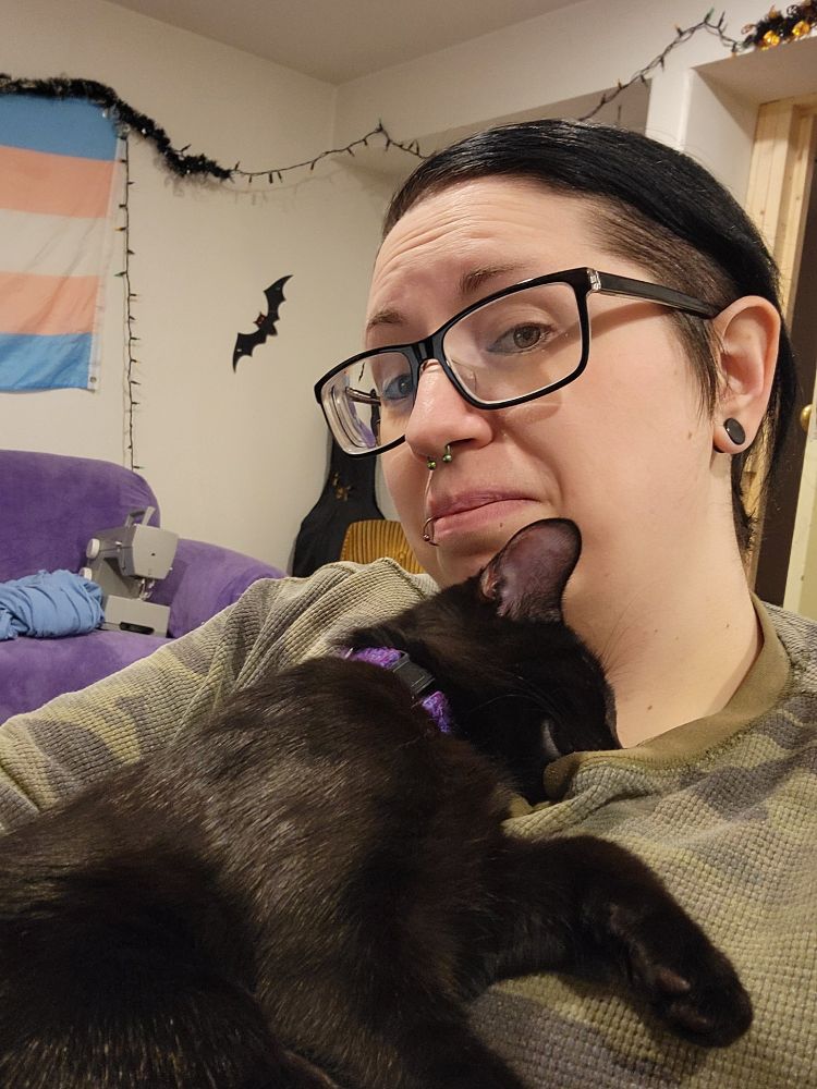 A human with dark hair, glasses, and piercings is almost tearfully happy while holding their 5 month old black kitten who is fast asleep on their chest. 