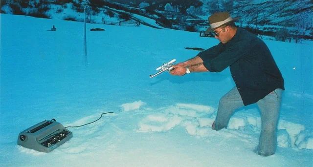 Hunter S. Thompson in Colorado. He is standing in the snow and pointing a handgun at his typewriter.