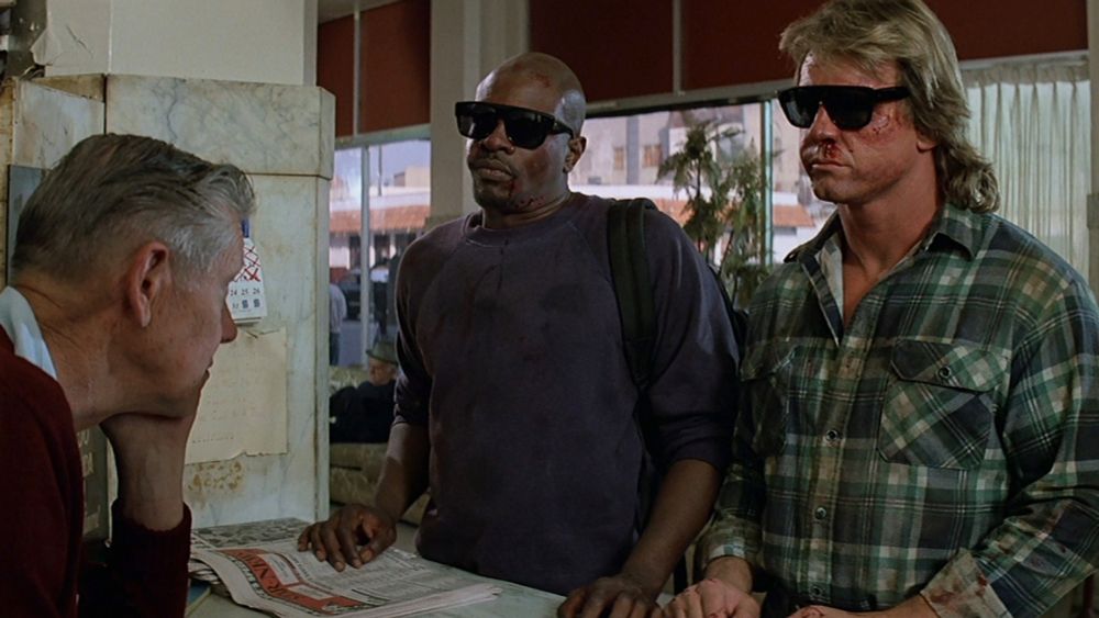 They Live. Keith David as Frank and Roddy Piper as Nada. They’re standing at a counter together. They both have sunglasses on.