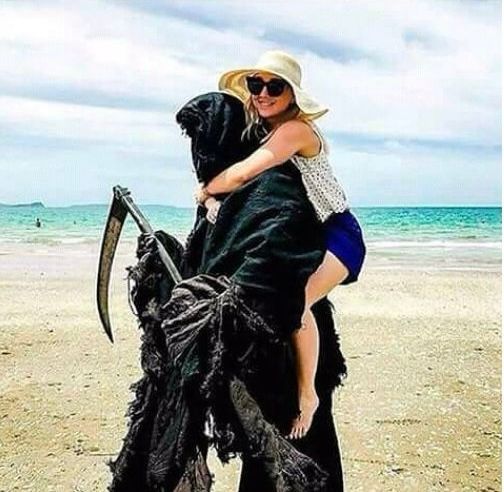 A woman getting a piggy-back ride from Death. They’re on a beach. It’s a sunny day. She has beach attire on. Death does not.