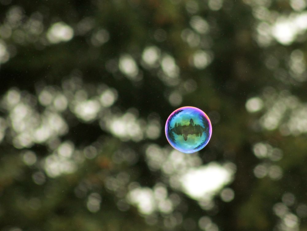 A single soap bubble floats in sharp focus against a blurred background of dark green trees. The bubble glows with iridescent colors—magenta, sapphire, and jade. At its center is a vivid reflection that resembles a silhouette of treetops curving around a central axis, almost like an inverted landscape inside a snow globe. The background is softly dappled with circular light spots from the bokeh effect, giving the image a dreamy, surreal atmosphere. This image is part of a series called DriftCodex, where each photograph captures ephemeral or mysterious moments, framed as interdimensional artifacts.