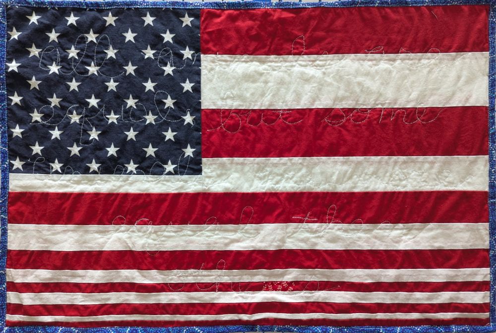 Protest sign: quilted United States flag pieced with uneven stripes. Words stitched into the quilt with white thread say "all animals are equal, but some animals are more equal than others"