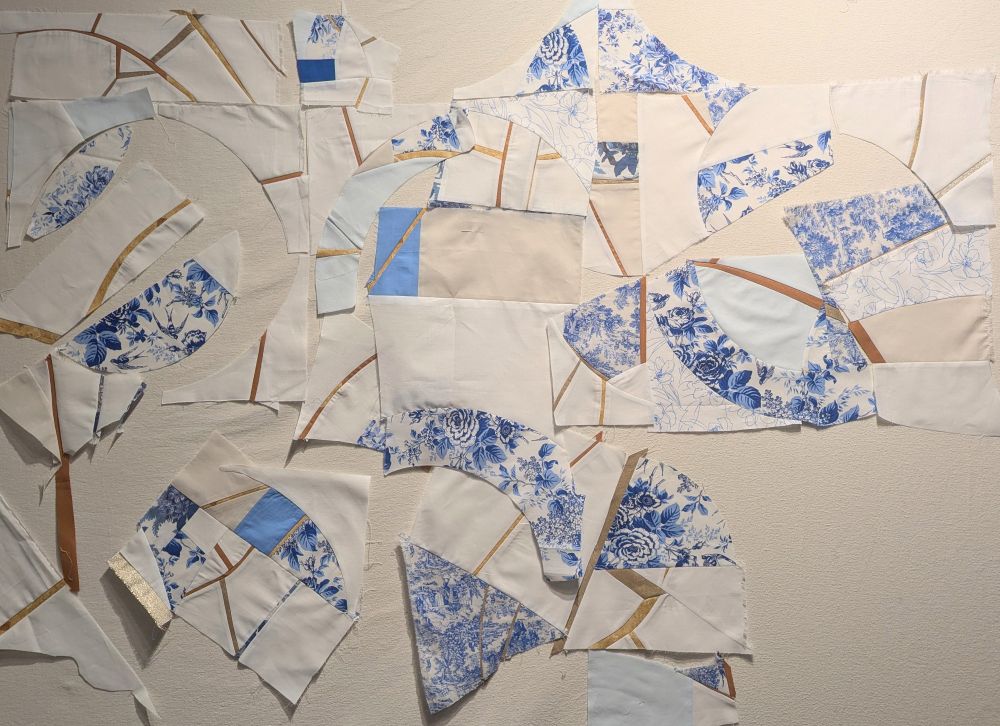 Scraps of blue, white, and gold fabric pieced into larger chunks and pinned onto a design wall
