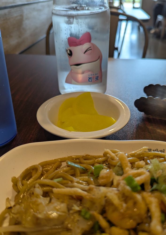 POV of somebody (me) sitting in front of a yummy plate of stir fried noodle. On the other side of the table is a plastic water bottle featuring a pink cartoon frog with a bow on its head blowing a kiss at...me? Oh wow oh gosh 