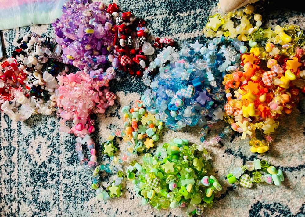 Piles of Kandi (beaded) bracelets sorted in different colors, (red and black and white combinations, purple, green, pink, blue, yellow, and orange) on a carpet.