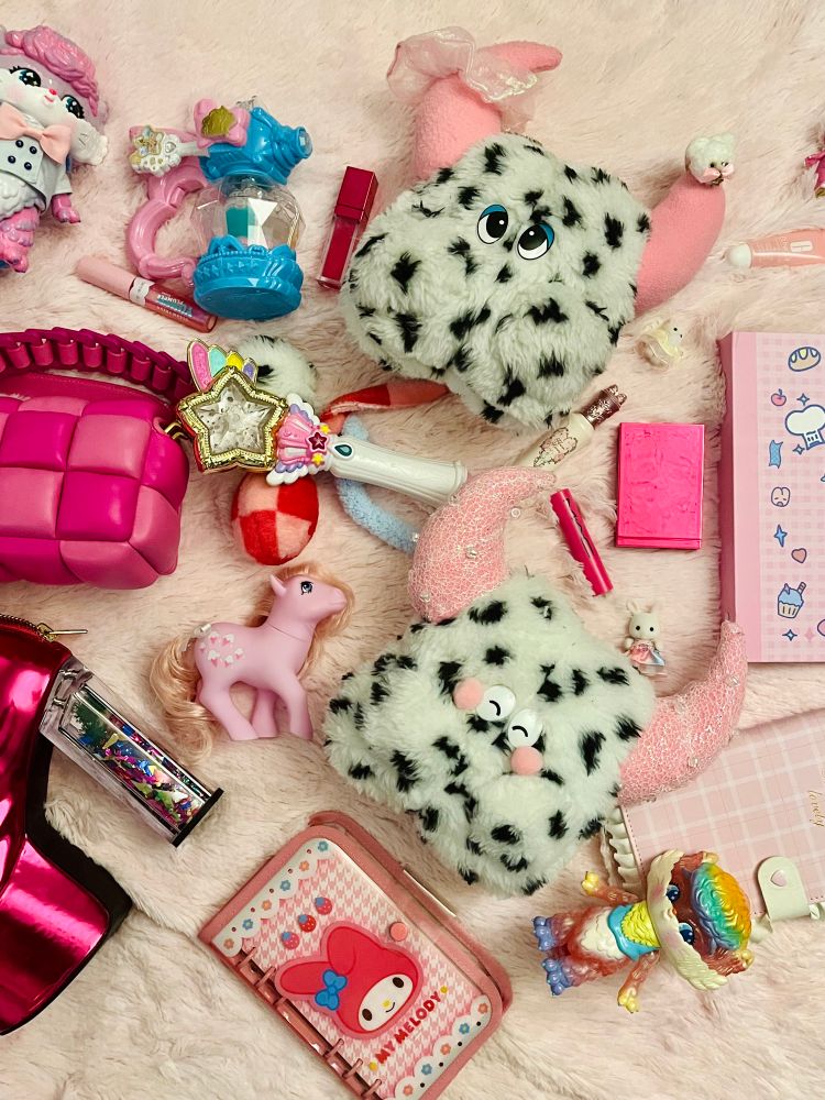 Two plush monsters with Dalmatian spots and pink horns lay on fluffy pink fabric surrounded by makeup, shoes, notebooks, toys, magical girl wands, and random girly objects. The monsters have long tails with funny orbs on the end in pink checkered and in blue. One is smiling with eyes closed and the other has her eyes open with blue eyeshadow in her eyelids. She has a pink iridescent scrunchie on one of her horns and a small bear on the other. The one with her eyes closed has lace and iridescent beading on her horns.