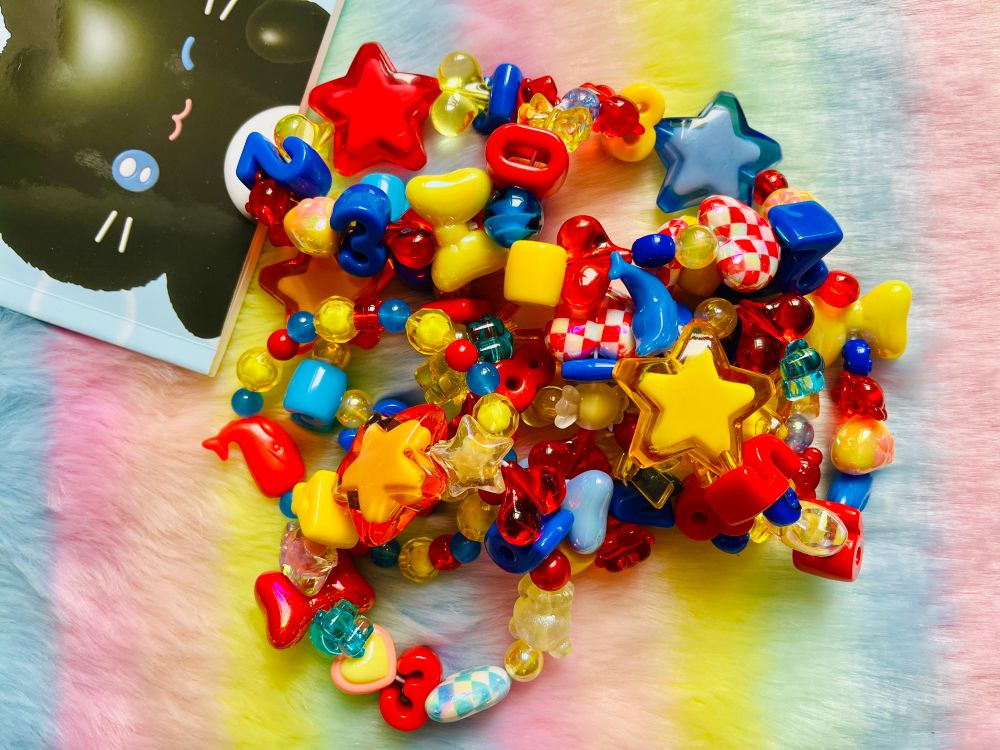A pile of primary colored Kandi (beaded) bracelets with chunky beads on a rainbow furry background, there is a cute cat notebook in the upper corner.