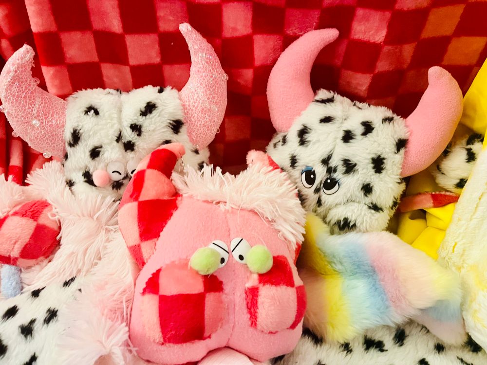 Three funny shaped monster plush in a pile of colorful fabric. The monsters are cute and colorful and have details such as lace, beading, pompoms, and checkers.