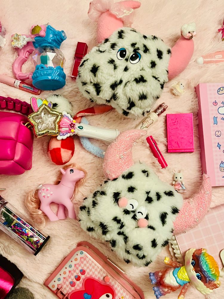 Two funny shaped fuzzy monster plush, they have Dalmatian spots and large pink horns. One of them has her eyes open and the other has her eyes closed with pompoms for blush. She has lace covered horns and iridescent beads on them. The other one has a pink scrunchie on one of her horns and a tiny bear on the other. They both have long tails with round orbs on the end. They are on a fluffy pink cloth, surrounded by cute toys, shoes, makeup, magical girl wands, and other girly items.