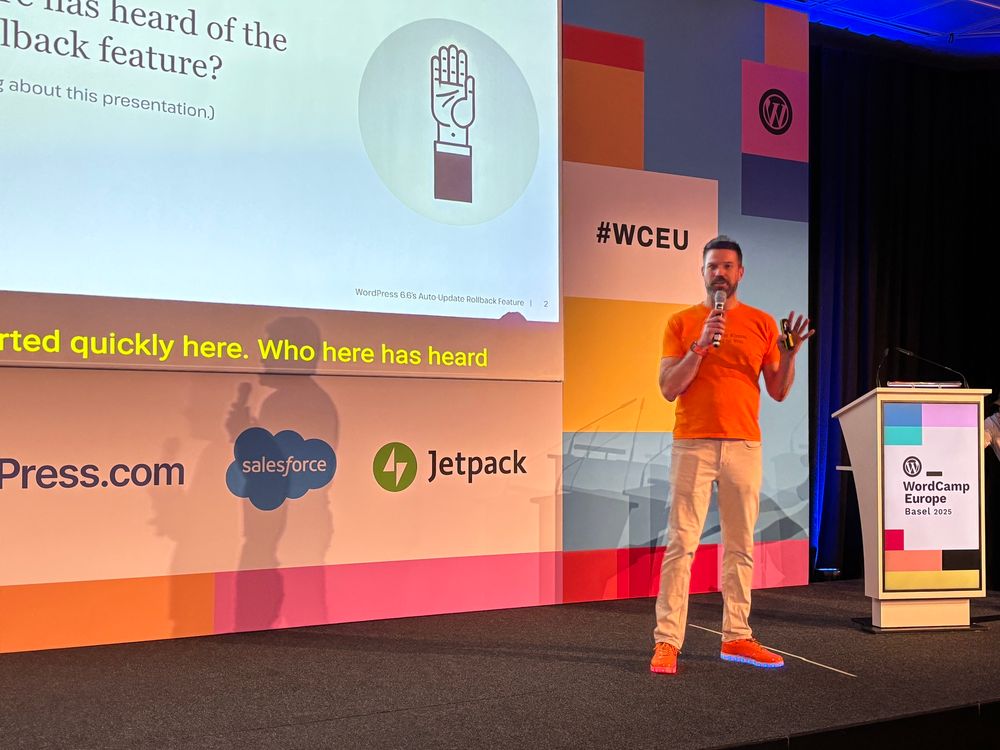 Roger Williams on stage with a microphone. He’s wearing an orange shirt and orange shoes with multi-colored lights in the soles. There’s a large screen showing a presentation behind him.  