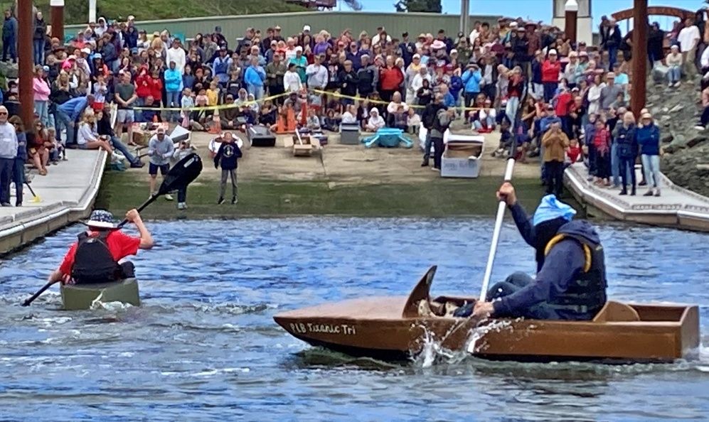 Cardboard boat races. 2 cardboard boats in the water, crowd looking on from boat launch 