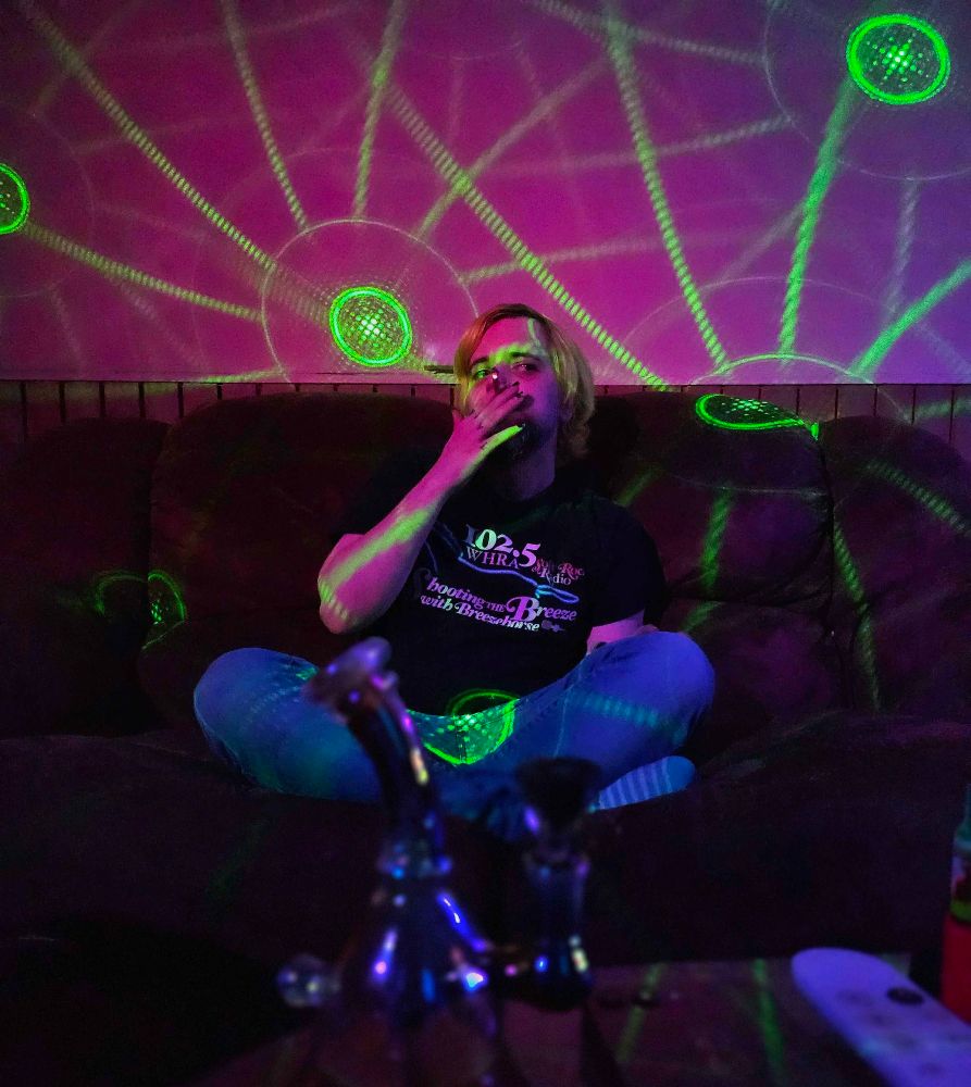 Photo of me sitting on the couch with my legs crossed, There are circular patterns made from a laser party light in the room. I'm smoking a joint