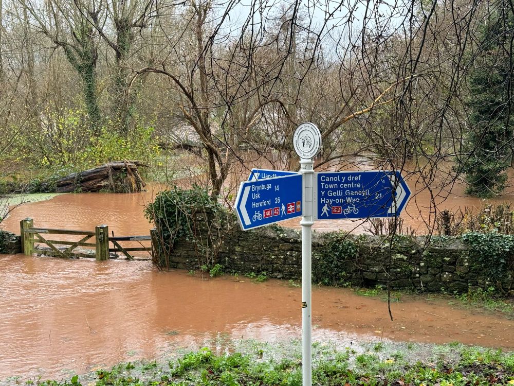 Dolydd y Castell, y Fenni, dan ddŵr yn llifo trwy ffensys. Topsoil-rich water flowing freely across the Castle Meadows, Abergavenny, with a cycle route sign in the foreground.