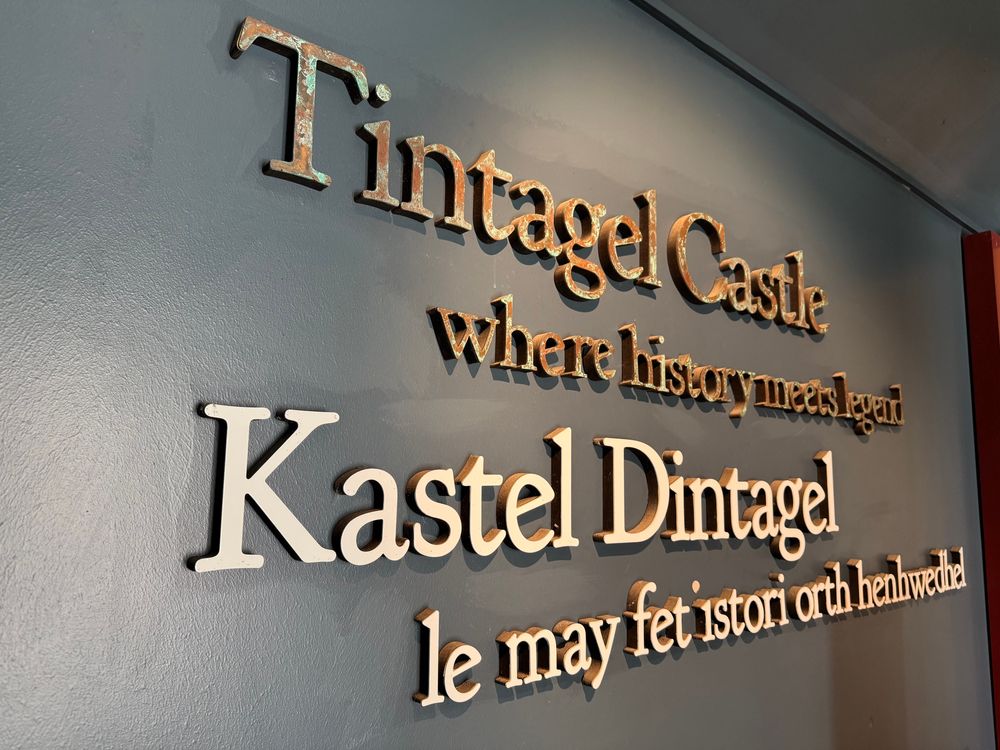 Wal gyda thestun metel yn Saesneg a Chernyweg wedi'i osod arni /  A wall with metal text mounted on it in both English and Cornish: “Tintagel Castle, where history meets legend - Kastel Dintagel, le may fet istori orth henhwedhel”