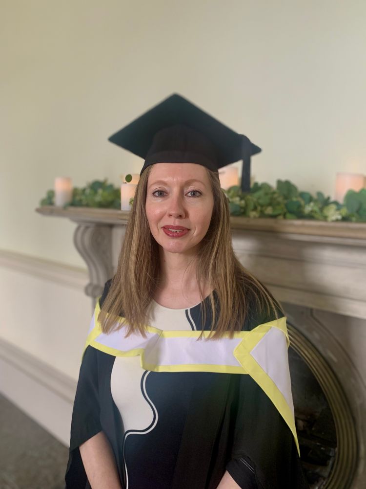 Picture of a woman in graduation robes standing in front of a period fireplace.