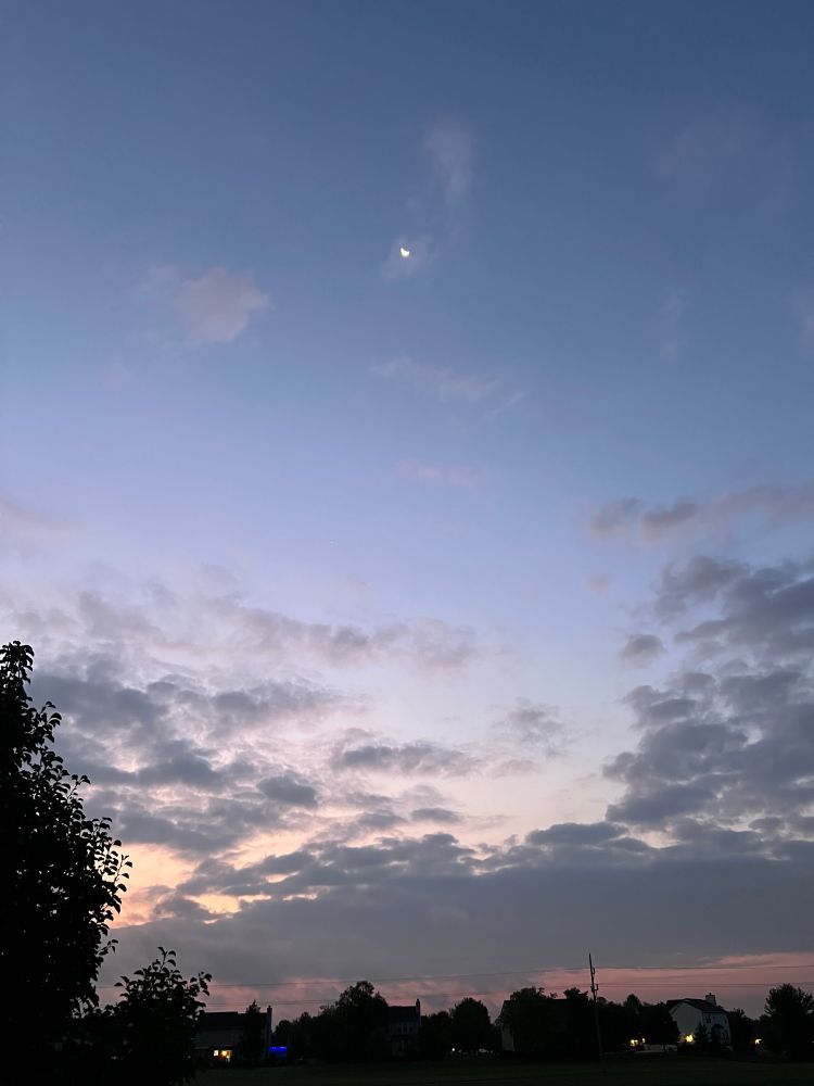 Grey clouds and peach sunrise beneath blue skies, moon sliver visible