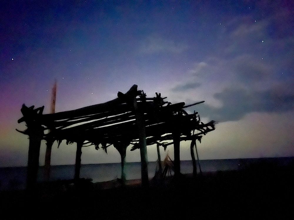 Wood structure in silhouette beneath a pink, yellow and green aurora sky