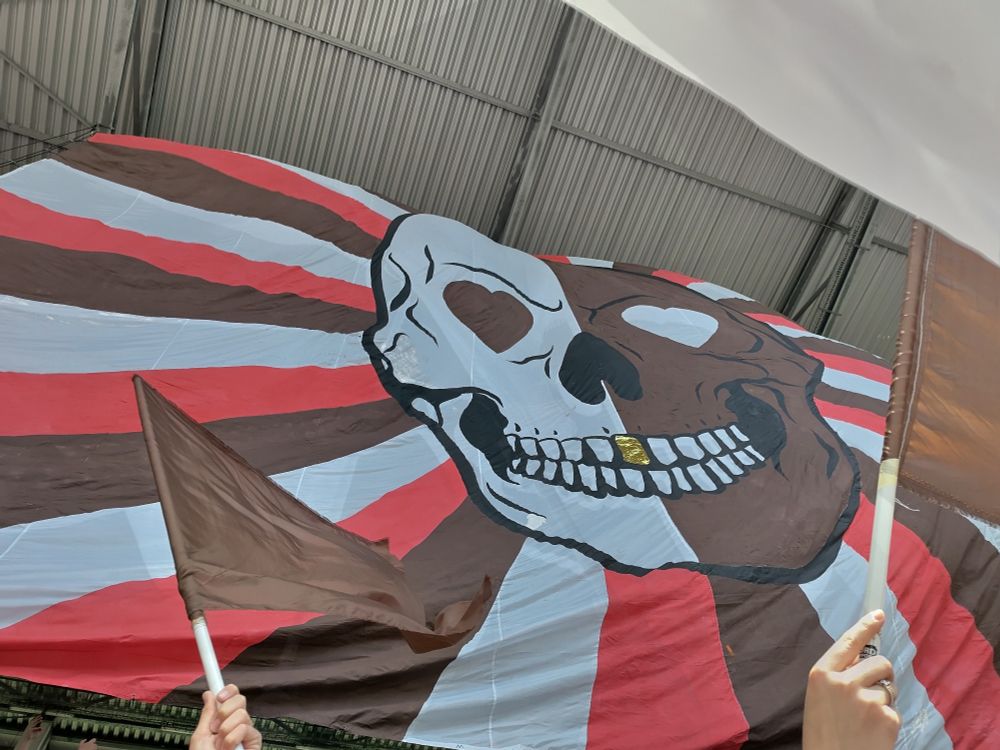 Die Totenkopf-Choreo von unten aus der Nordkurve fotografiert: der zentrale Totenkopf vertikal getrennt links weiß rechts braun, die Augen jeweils in der anderen Farbe als Herz. Hinter dem Totenkopf gehen braun-weiß-rote Strahlen aus in alle Richtungen. Das i-Tüpfelchen ist in der Mitte des Oberkiefers ein Goldzahn