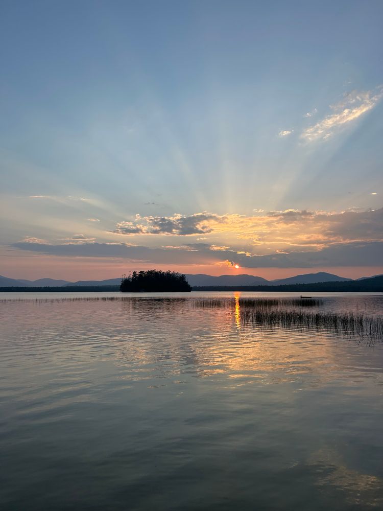 Sunset on a lake in Maine. The orange sun throws beams across the sky over purple mountains. It’s all reflected in silver water. There’s a black island in the centre of the lake