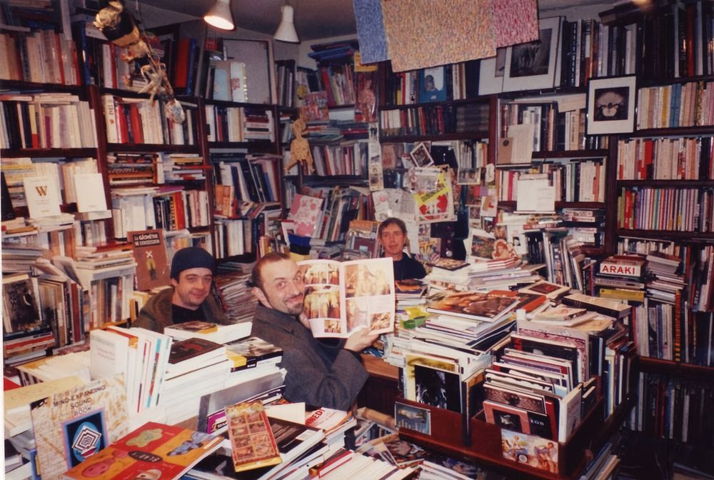 De gauche à droite : Kerozen, Pakito Bolino et Jacques Noël à la librairie Un Regard Moderne, circa 1997, droits réservés.
Trois personne dans une librairie. La quantité et la densité de livres est telle qu'on croirait qu'ils s'y baignent. Seules leurs têtes et leurs épaules dépassent des piles. Ils sourient. 