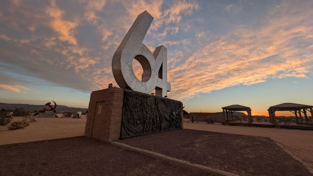 a giant sculpture of the number 64, with a relief below. It mashing up the US Civil War, World War II, and Tiananmen Square

in the distance, a hammer and sickle rests on a pyramid of skulls 