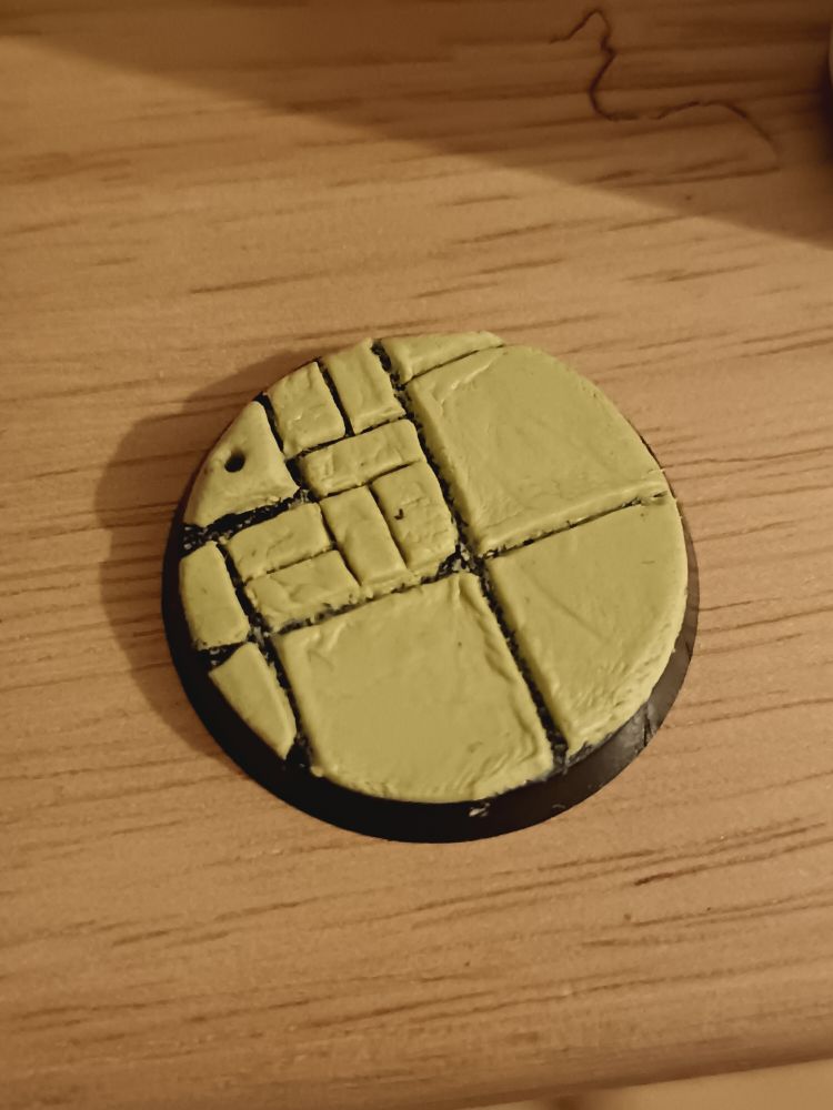 Top down pic of a warhammer base with dull yellow epoxy on it sculpted into brickwork, one corner is a short mound with a hole in it.