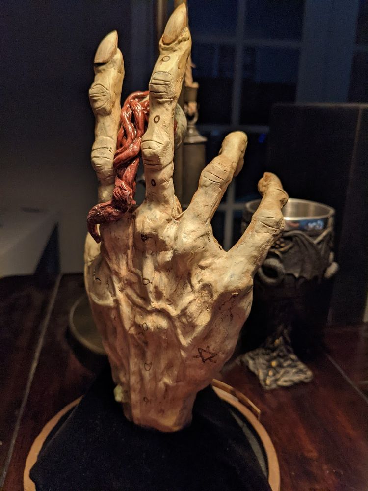 The back of a desiccated hand holding a yellow/orange eye. The hand has runes engraved in the skin. The back of the eyeball has shiny red nerve endings hanging down the back of the hand.
