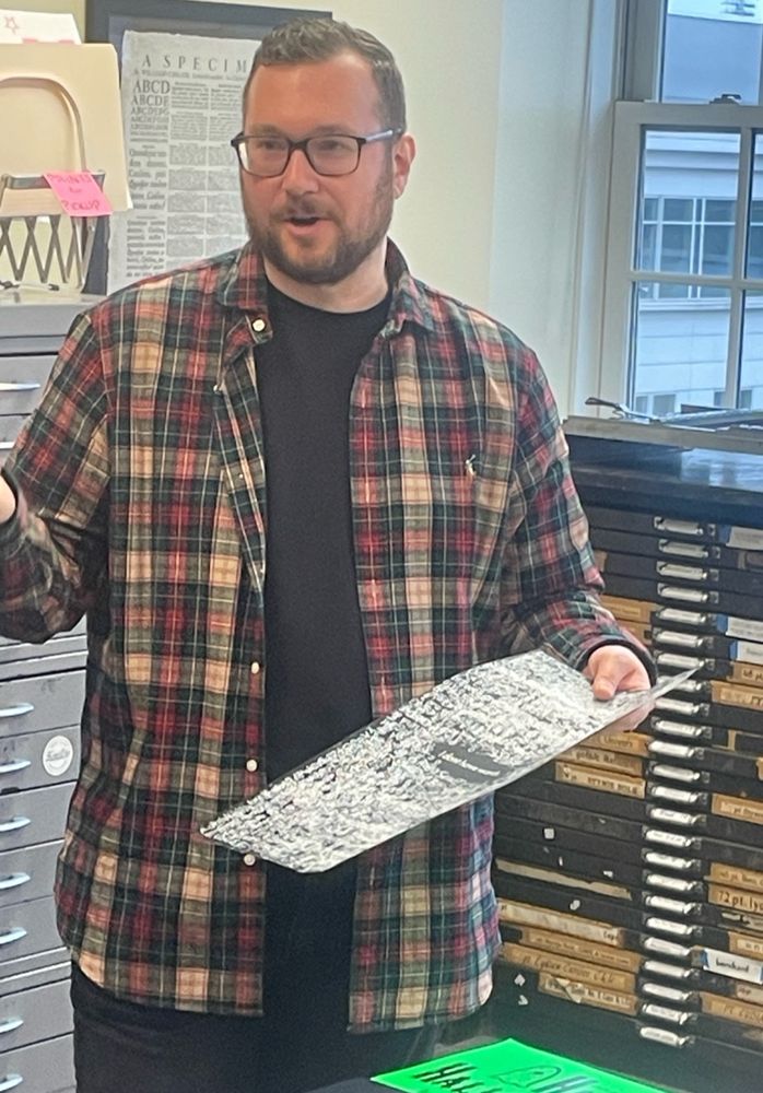 Photo of a man wearing a plaid shirt holding up a single sheet of paper with numerous letterpress ink impressions on it