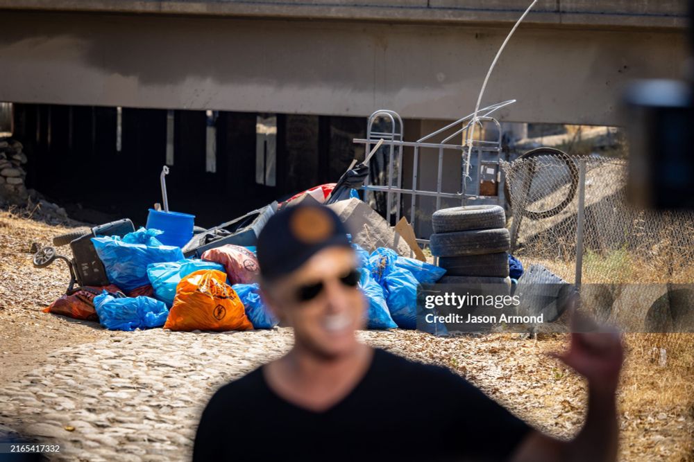 Gavin Newsom, out of focus, smiling and raising his hand with the bagged-up results of his camp sweep in clear focus behind him