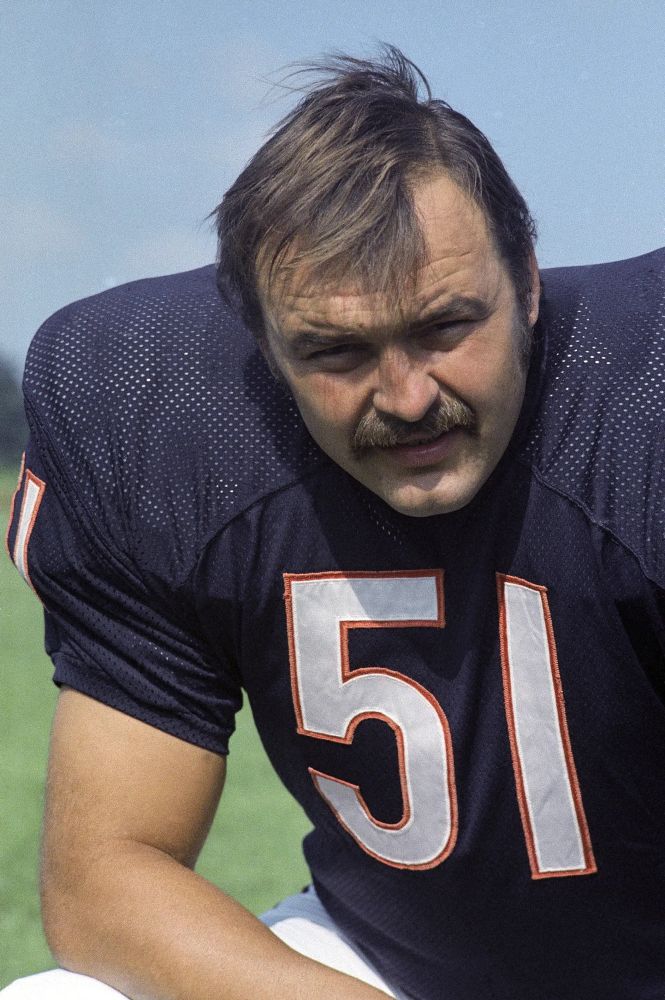 Dick Butkus kneeling with his arm on his leg. He’s wearing a blue Chicago Bears jersey with a white number 51 with orange trim around the numbers. 