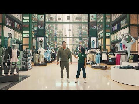 A man in grey sweats standing next to a shorter woman with dark hair wearing jean capris and a Dicks Sporting Goods polo. They are in the middle of a sporting goods store with their backs to the entrance. 