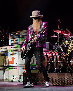 Billy Gibbons of ZZ Top playing a Gibson SG and wearing a fuschia and black/white tiger striped jacket, black pants, silver shoes, and his signature sunglasses. He is standing in front of a drum set with a stack of multi color painted amplifiers beside it. 