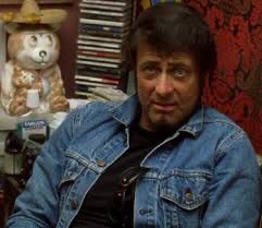 A man in a blue denim jacket, with a disbelieving look in his face, sitting behind a desk. Larry Gomez is a character in Kill Bill