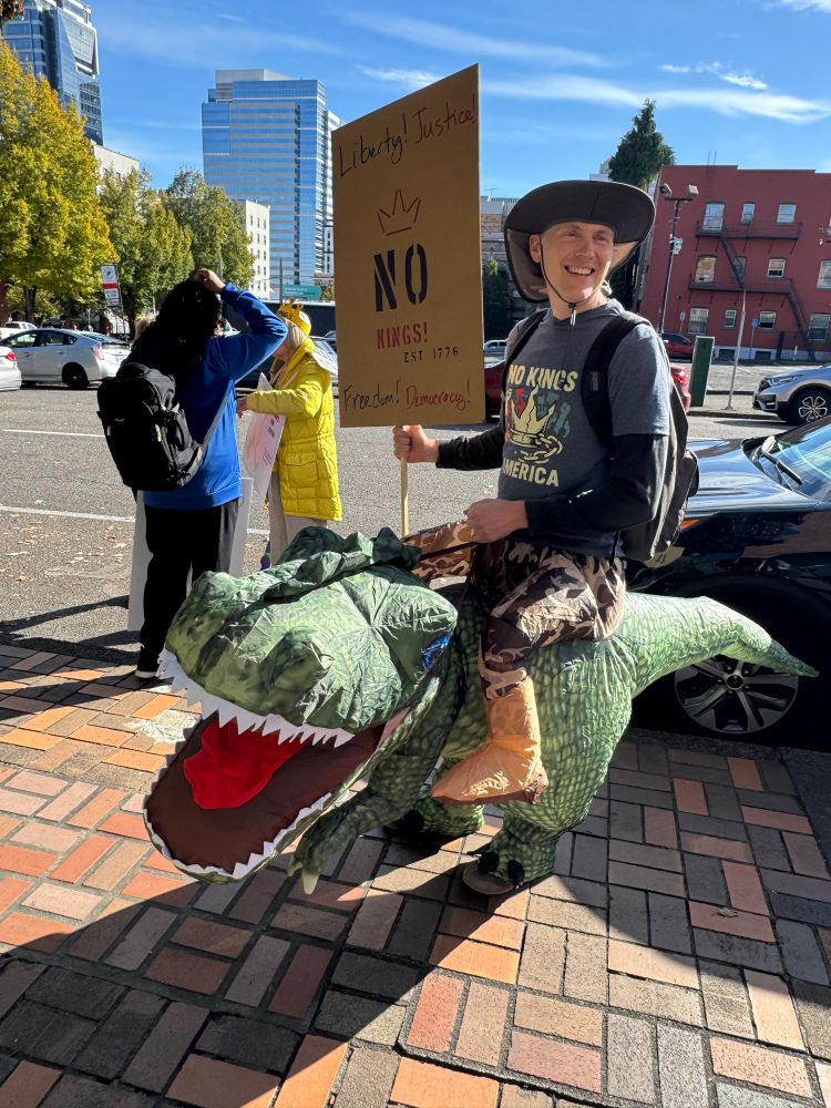 Man marching with First Unitarian Church, #Portland on No Kings day . riding on an inflatable dinosaur costume, posing for photo. 