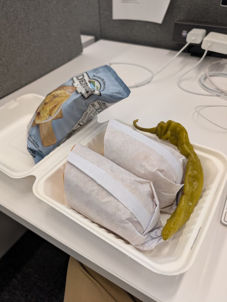 On a desk, a bag of chips sits inside one half of a paperboard deli box. In the other half is a hero wrapped in paper and split in half. Tucked around the sandwich is an absolutely enormous pickled long pepper. It wraps around one side of the container and most of another.