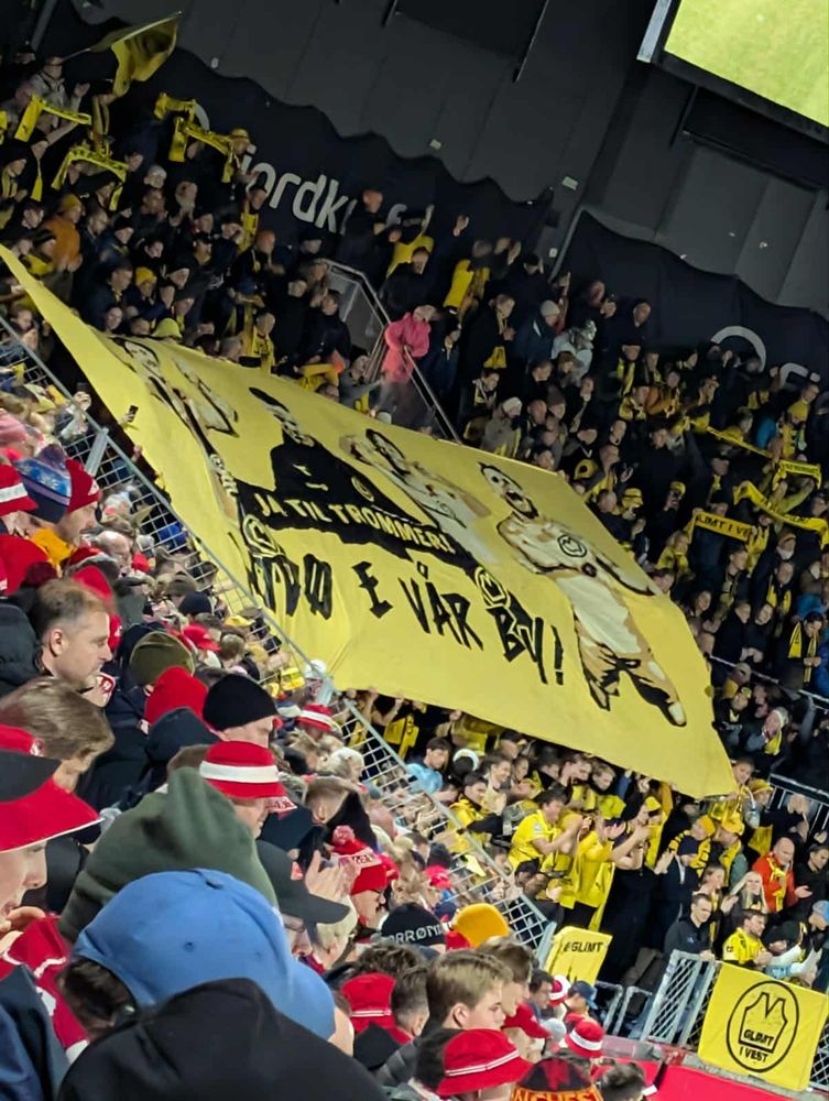 Bilde av bortefeltet til Bodø/Glimt mot brann med TIFO. TIFO består av Moberg, Knutsen, Mvuka og Blomberg med teksten "Bodø e vår by". Knutsen holder et skjerf der det står "Ja til trommer"