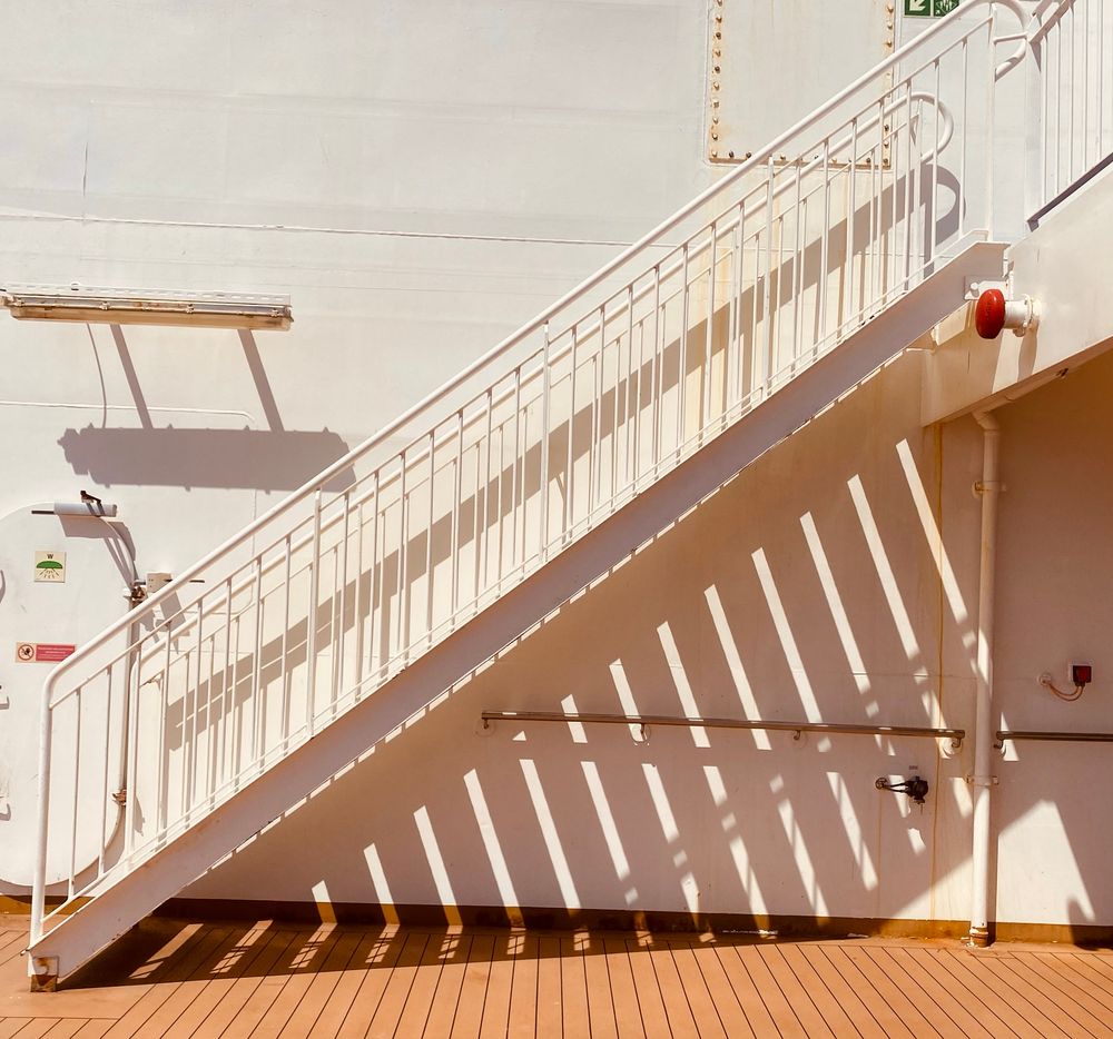 A staircase on the deck of a passenger ferry in bright sunshine, which creates striking shadows on the wall behind and the floor below. 