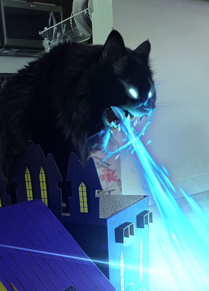 A black cat standing on a spooky castle-like model building. Its mouth is open wide and face twisted into a scream. The photo has been edited to show it shooting a blue flame from its mouth like Godzilla.