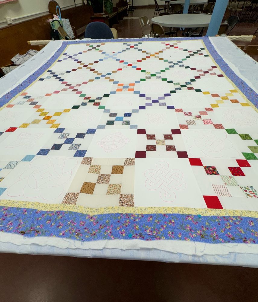 Almost queen size hand sewn quilt in a hand quilting frame. It is made from scraps of many colored fabrics alternating with white blocks. The pattern is called Irish Chain