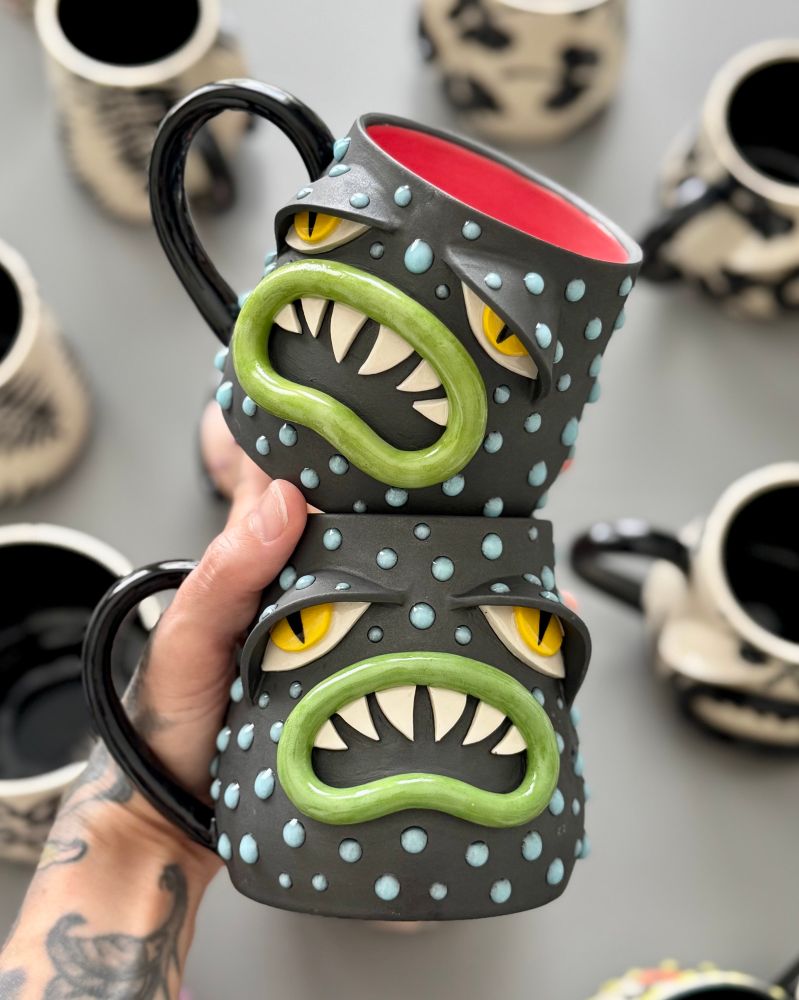 A photo of two Sad Boi mugs, painted with a variety of underglazes. The mugs have sculpted facial features including lips with sharp teeth, and eyes with exaggerated lids. The body of the mug is black with textural blue gloops. 
