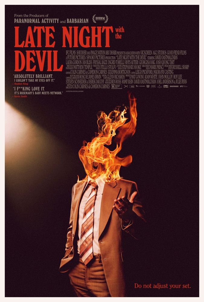 Theatrical poster for the Shudder original film "Late Night with the Devil."

The poster shows a man in a brown suit, standing against a black background with a spotlight to his right. His right hand is in his pocket, while his left hand is raised. His head is on fire, with only a screaming mouth visible.